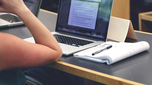 A black desk with a laptop open with a notepad and pen next to it. A person sits with their elbow in the table, looking at the screen.
