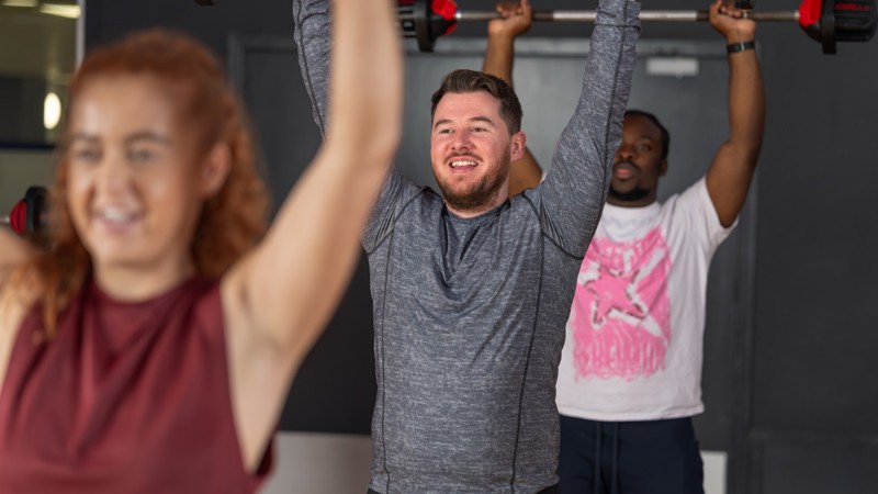 Three people smiling during a fitness class, all of them are holding barbell weights above their heads.