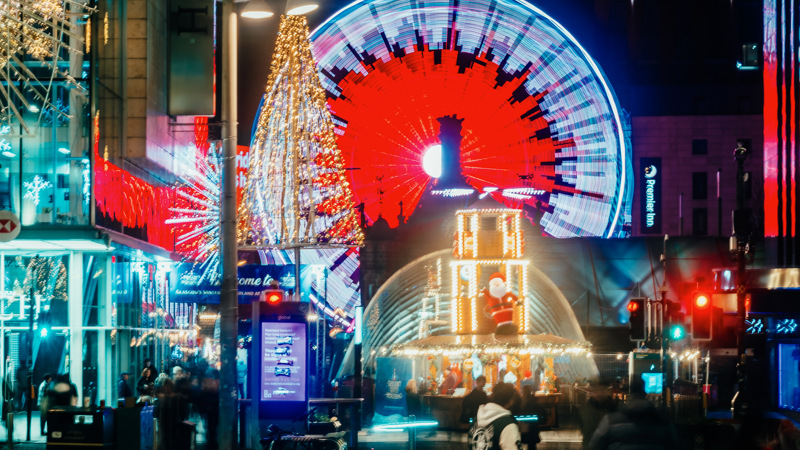 A vibrant city street at night decorated with Christmas lights, featuring a large illuminated Ferris wheel in the background, a glowing Christmas tree-shaped installation, and a carousel surrounded by festive lights and people walking.