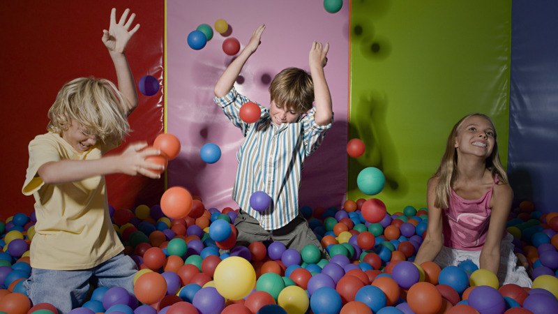 Three children play in a colourful soft‑play ball pit, tossing bright plastic balls into the air. They are surrounded by multicoloured padded walls, and the scene is full of movement and energy as they enjoy the activity.