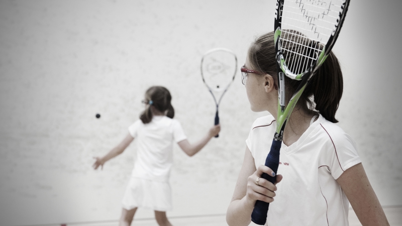 Two young players wearing white sports outfits are on a squash court. One stands in the foreground holding a racquet and watching the ball, while the other is positioned closer to the wall, swinging their racquet to return the shot.