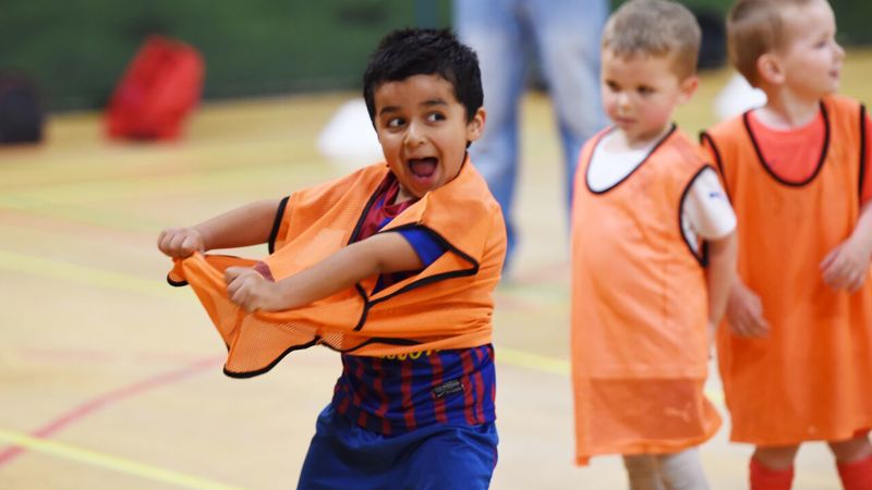 Children wearing orange sports bibs participate in an indoor activity. One child in the foreground is lifting their bib over their head while others stand nearby, waiting and watching. The scene takes place in a gym with a wooden floor and sports cones in the background.