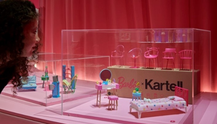  A woman leans in to view a glass display case containing miniature Barbie-branded furniture and accessories, including chairs, lamps, and a bed, set against a backdrop of pink curtains.